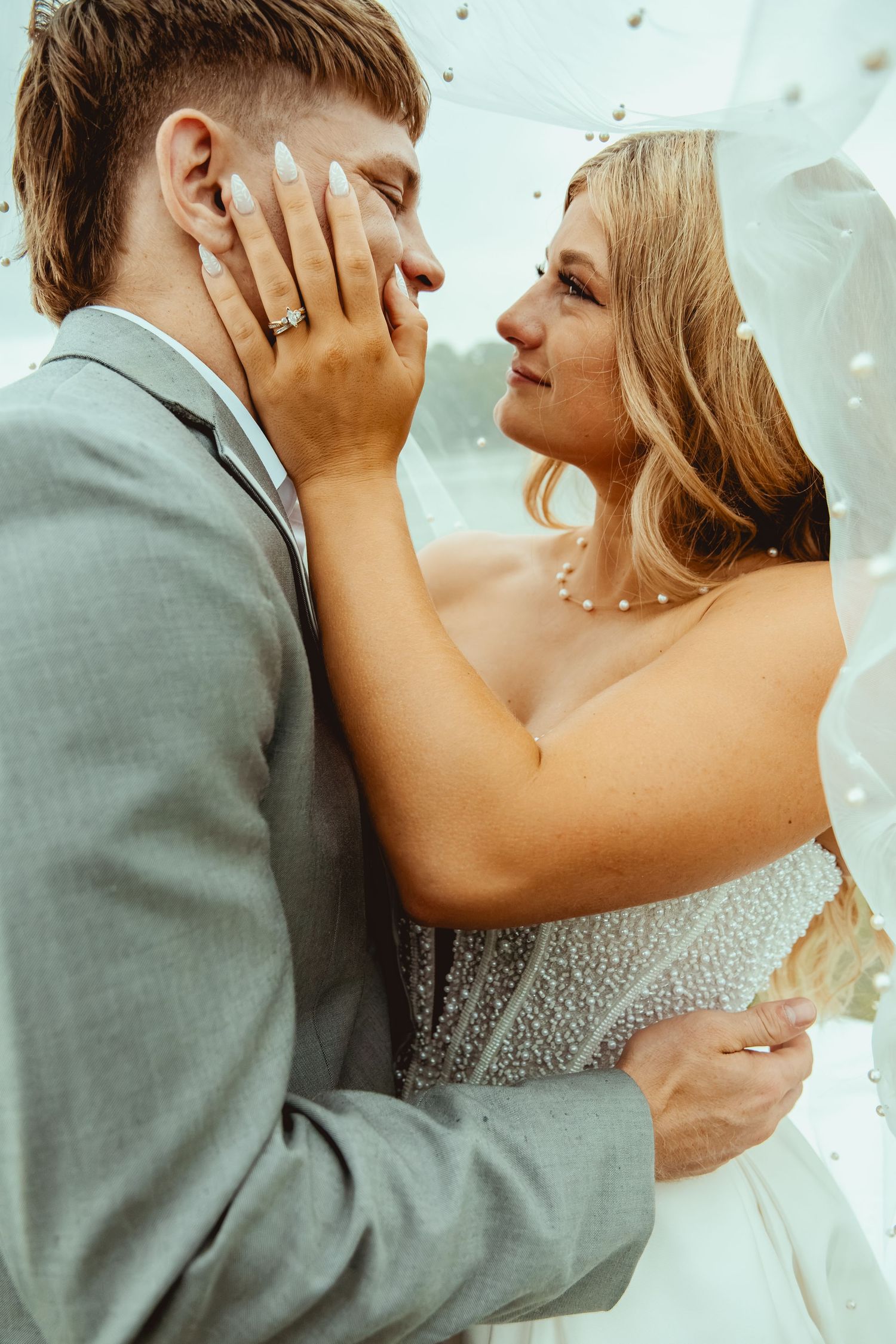 Bride tenderly holds grooms face under a veil, both gazing at each other in wedding attire.