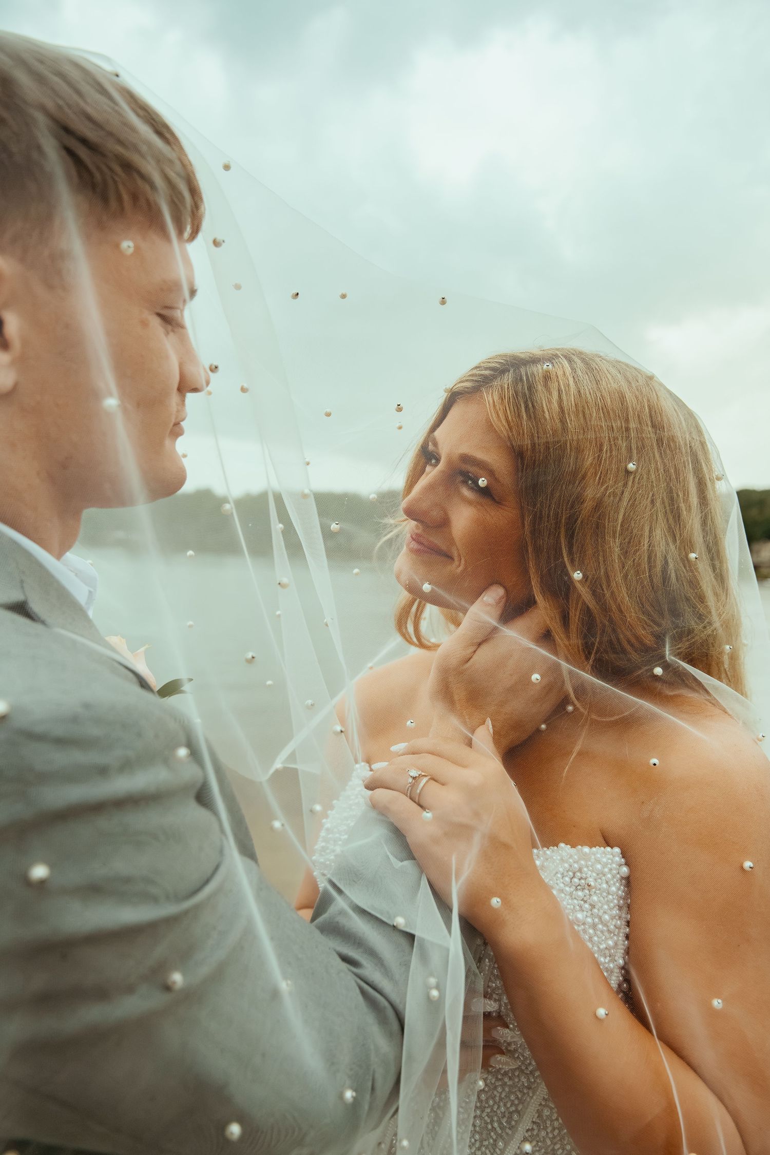Bride and groom share an intimate moment under a pearl-dotted veil, with the bride wearing a diamond ring.