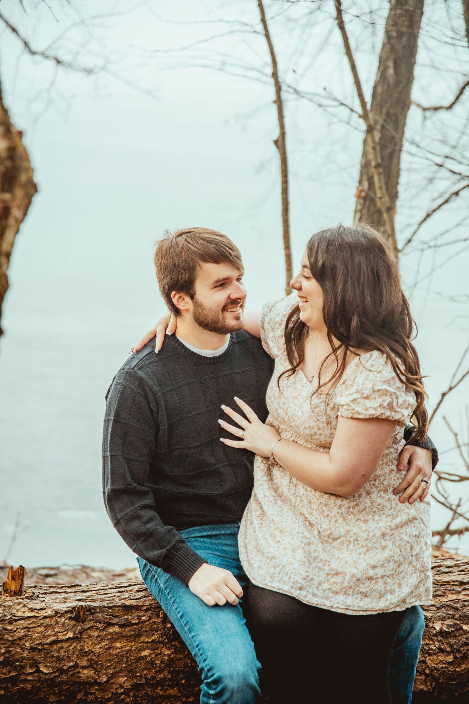 A couple sits together on a fallen tree trunk in a winter forest setting sharing an intimate moment.