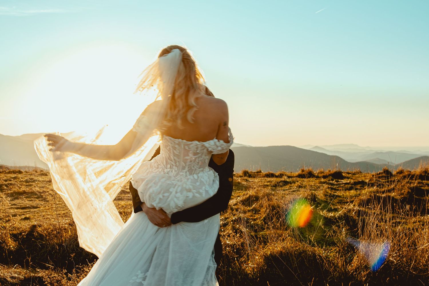 Bride and groom share intimate moment at sunset with flowing white dress and mountain landscape.