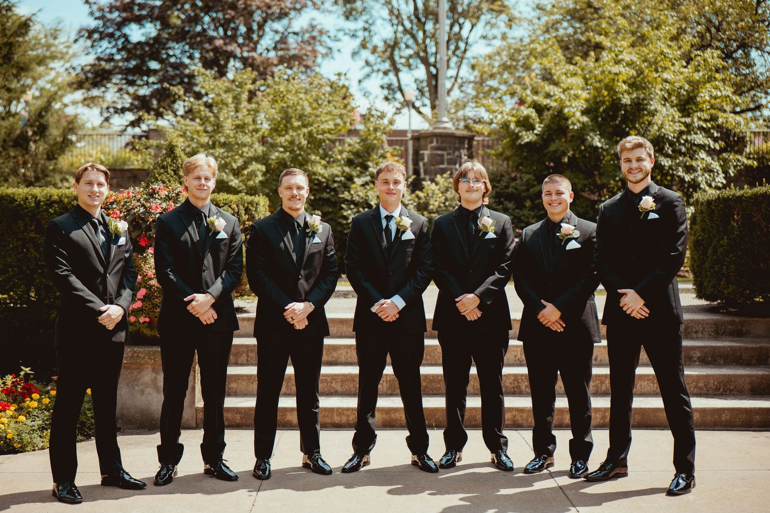 A group of groomsmen in black suits and ties stand in a line outdoors during a wedding ceremony.