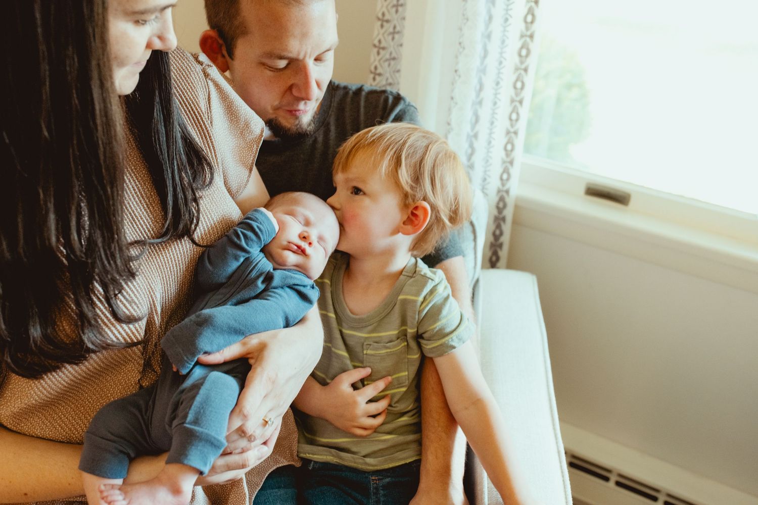 A family sits together near a window, with a toddler in green and an infant in blue.