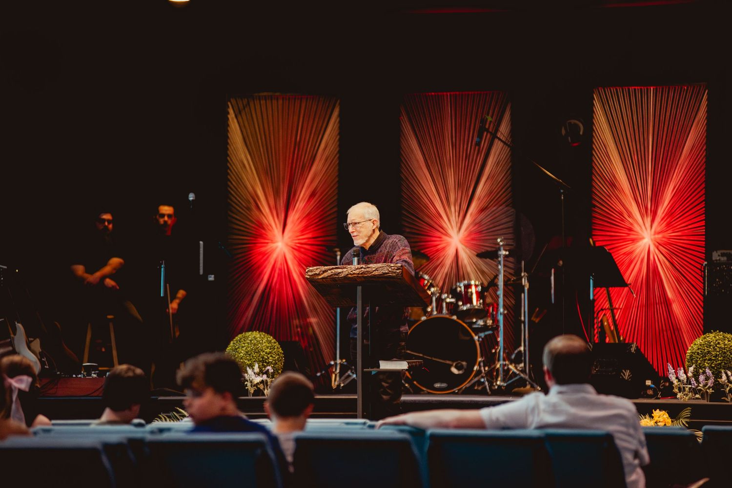 A performer stands at a podium on stage with dramatic red lighting effects and beams creating a striking backdrop.