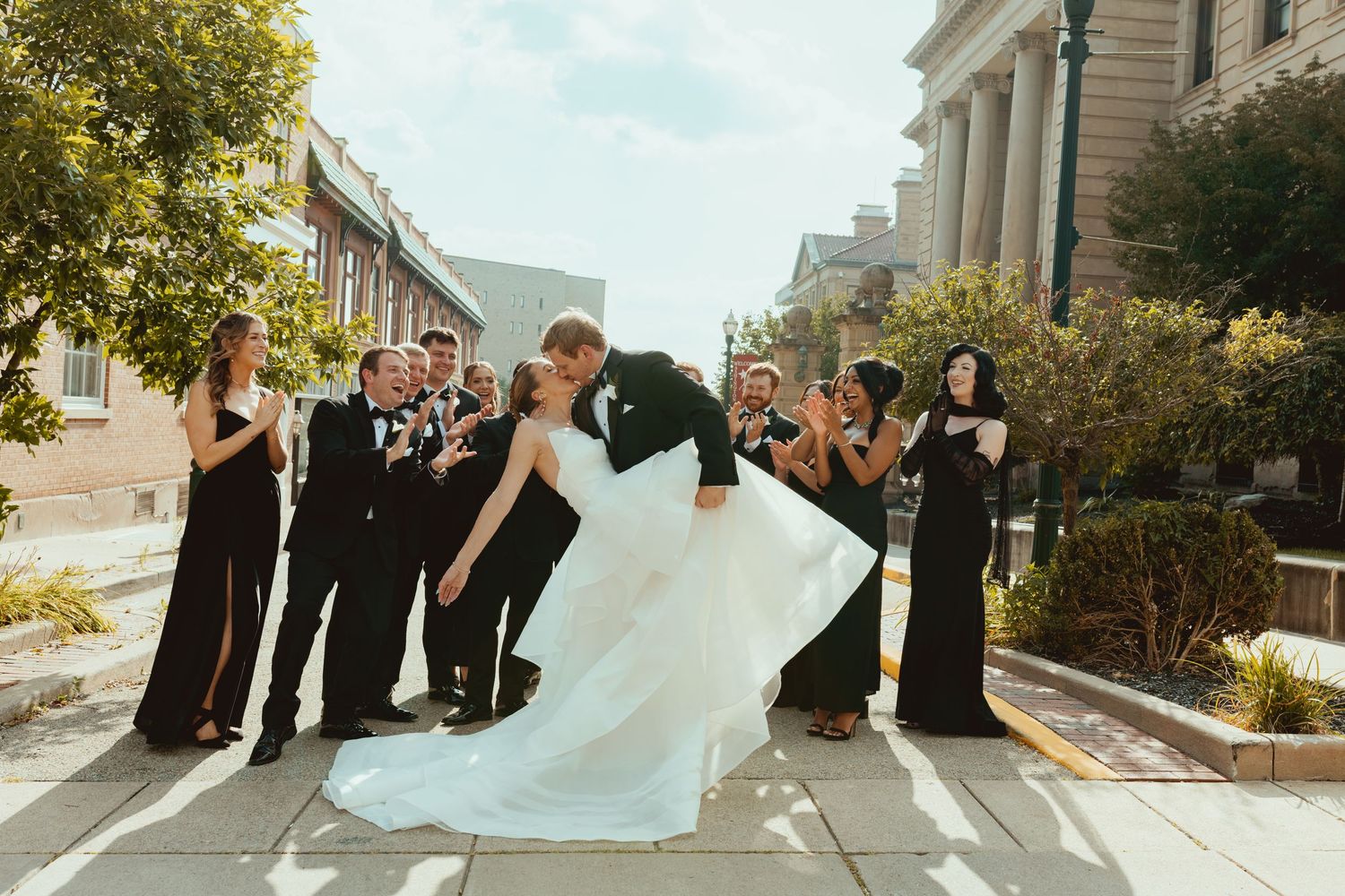 Wedding party poses for a fun photo on a sunny street with historic buildings in the background.