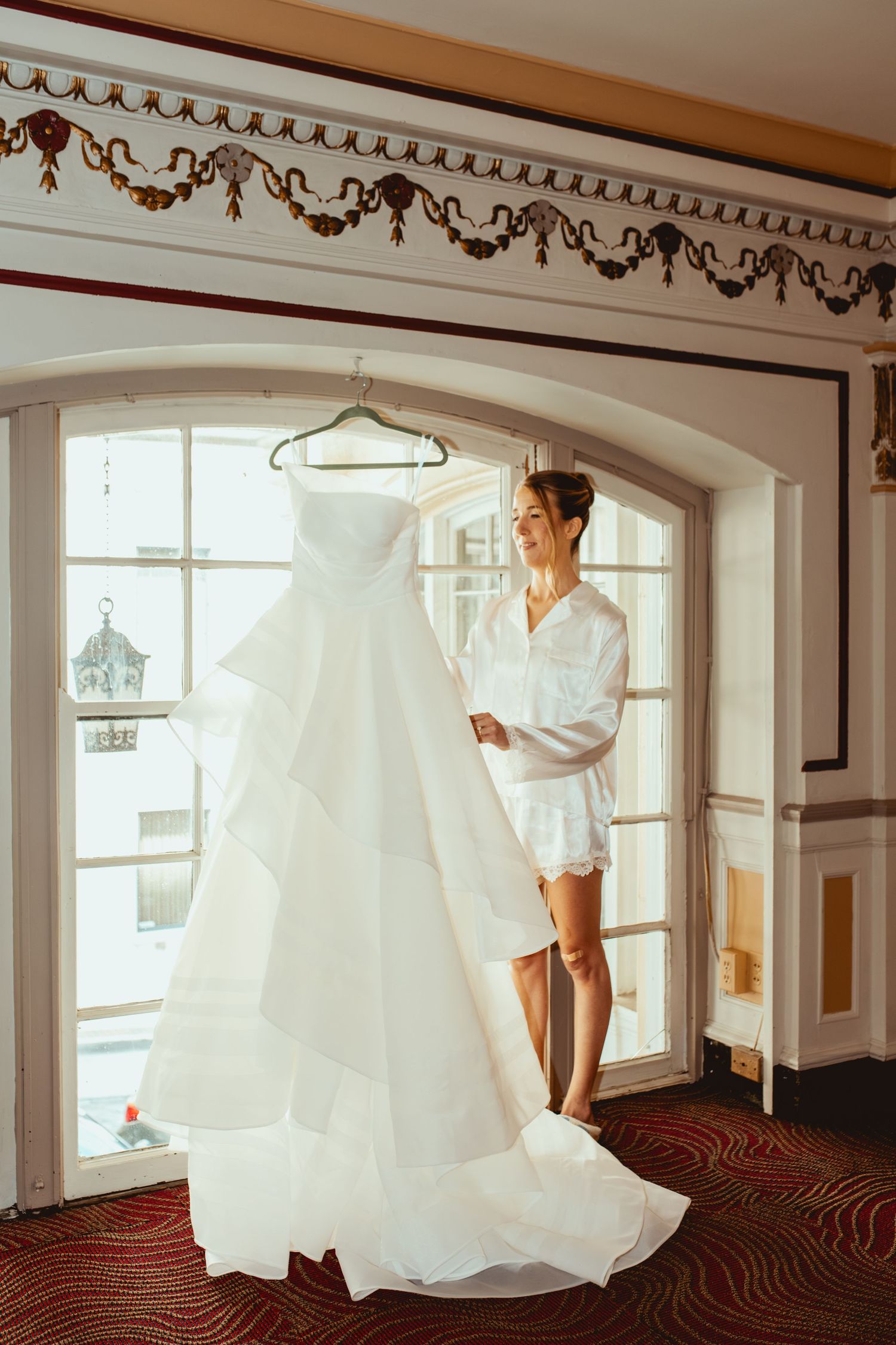 A wedding dress hangs in an elegant room with ornate crown molding and large windows.