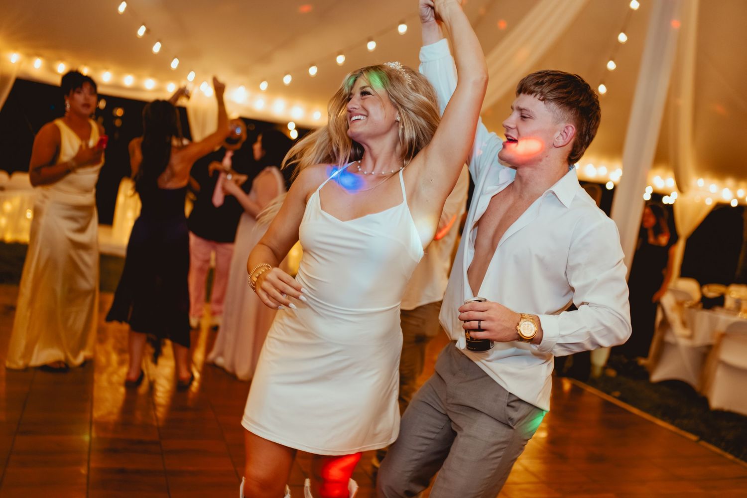 Bride and groom dance joyfully at lively wedding reception under string lights.