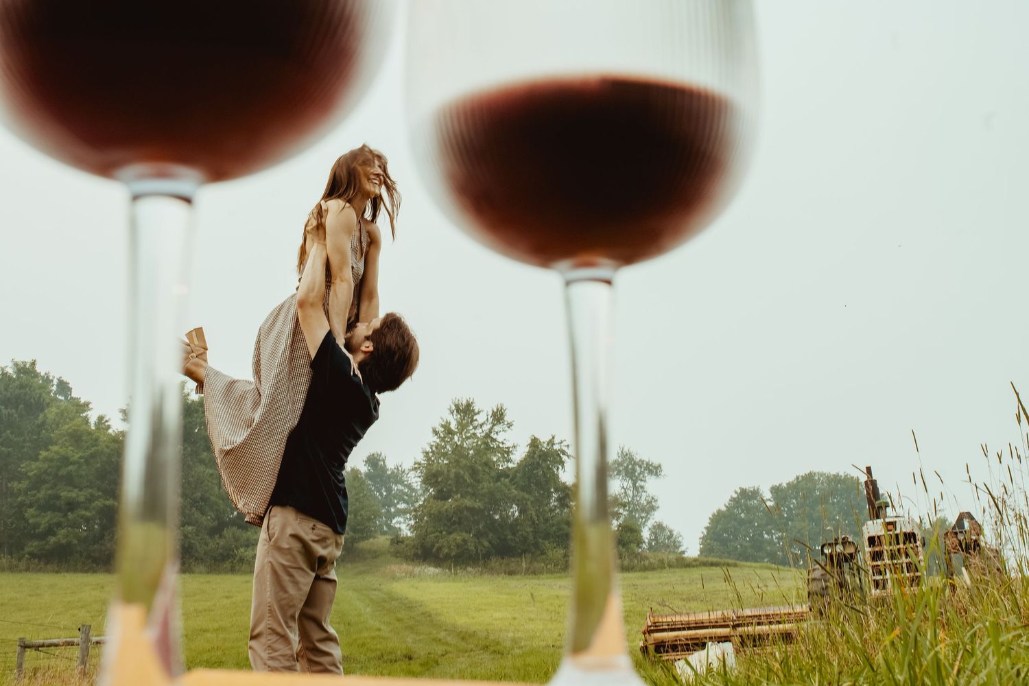 Romantic couple dancing outdoors at a vineyard with two glasses of red wine in the foreground.