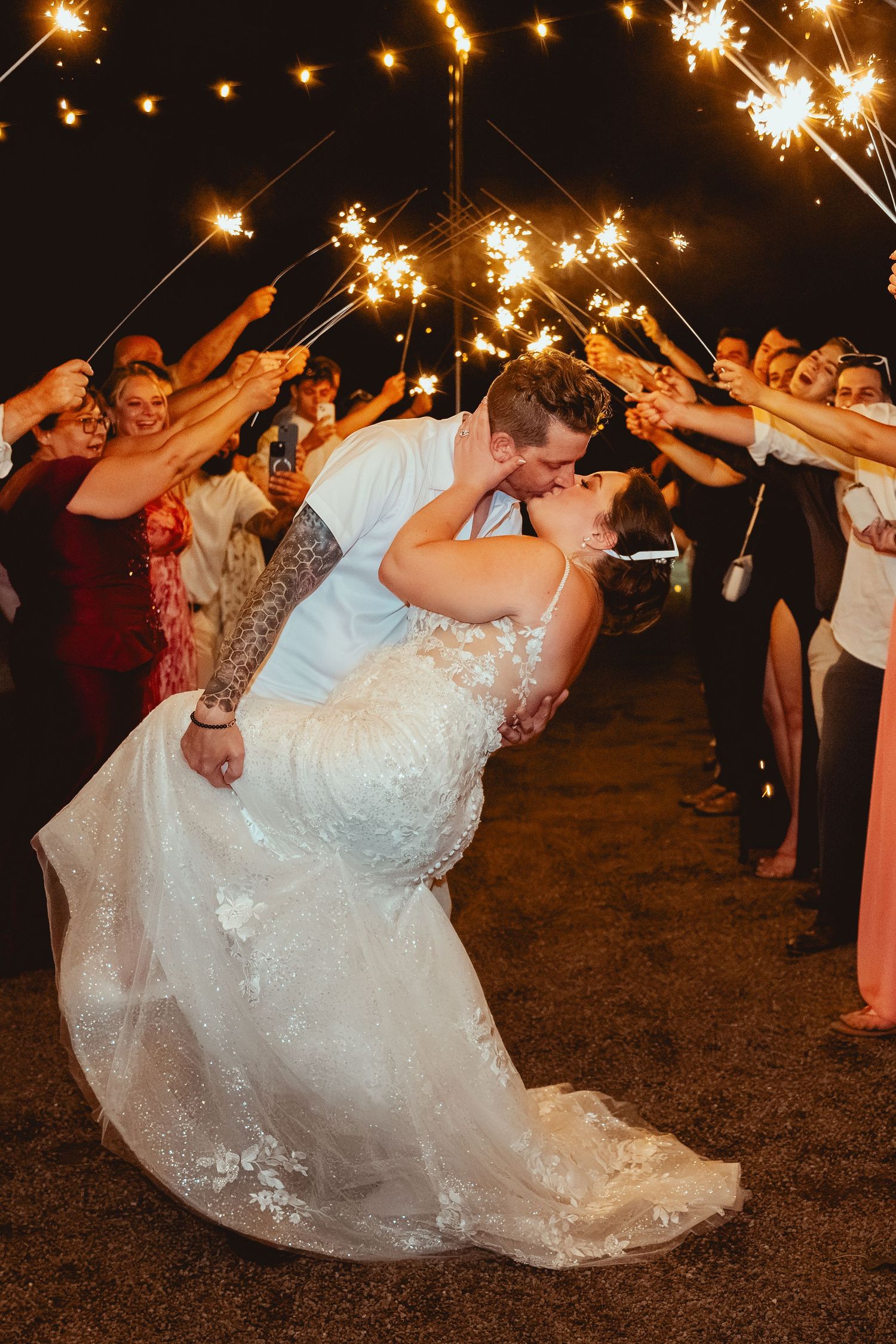 Bride and groom share a romantic dip kiss during a sparkler send-off surrounded by cheering wedding guests at night.