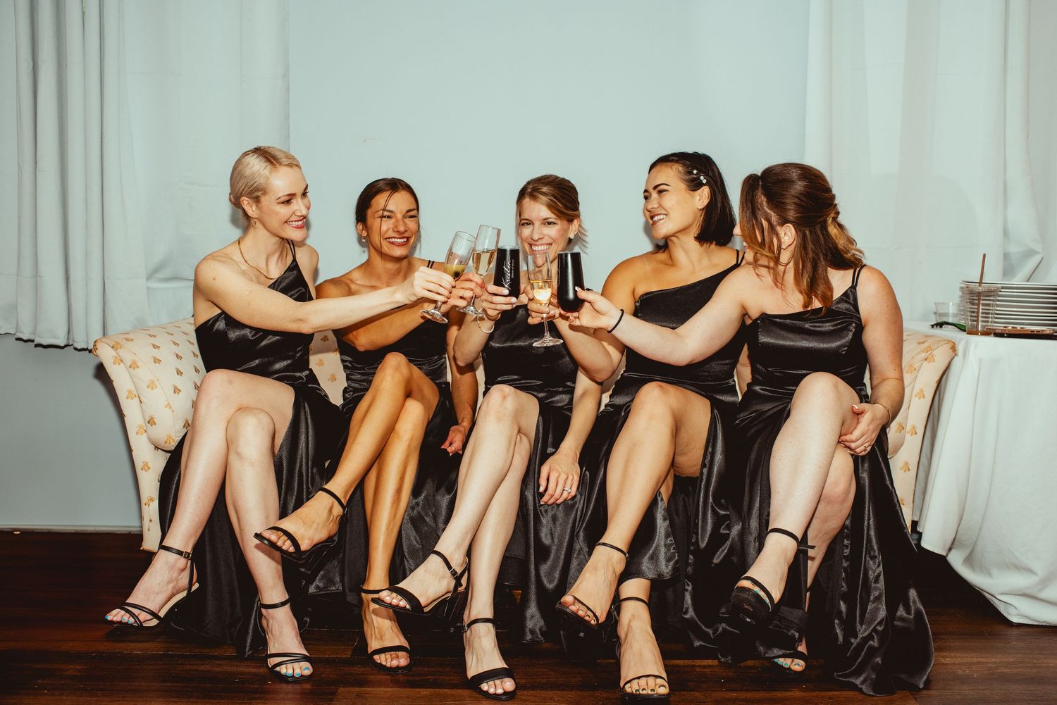 A group sits together on a black leather couch in a casual setting with white curtains in the background.