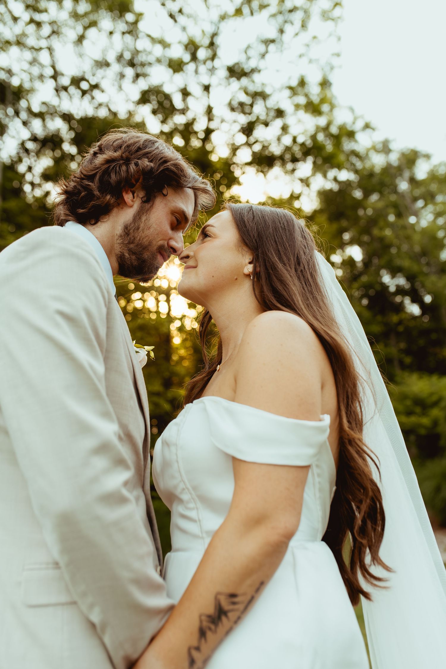 Bride and groom share a romantic forehead touch at golden hour surrounded by green trees.