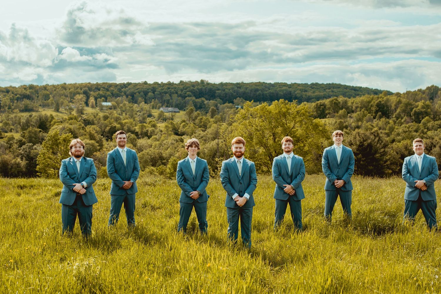 Groomsmen in matching teal suits standing in formation on hilltop meadow.