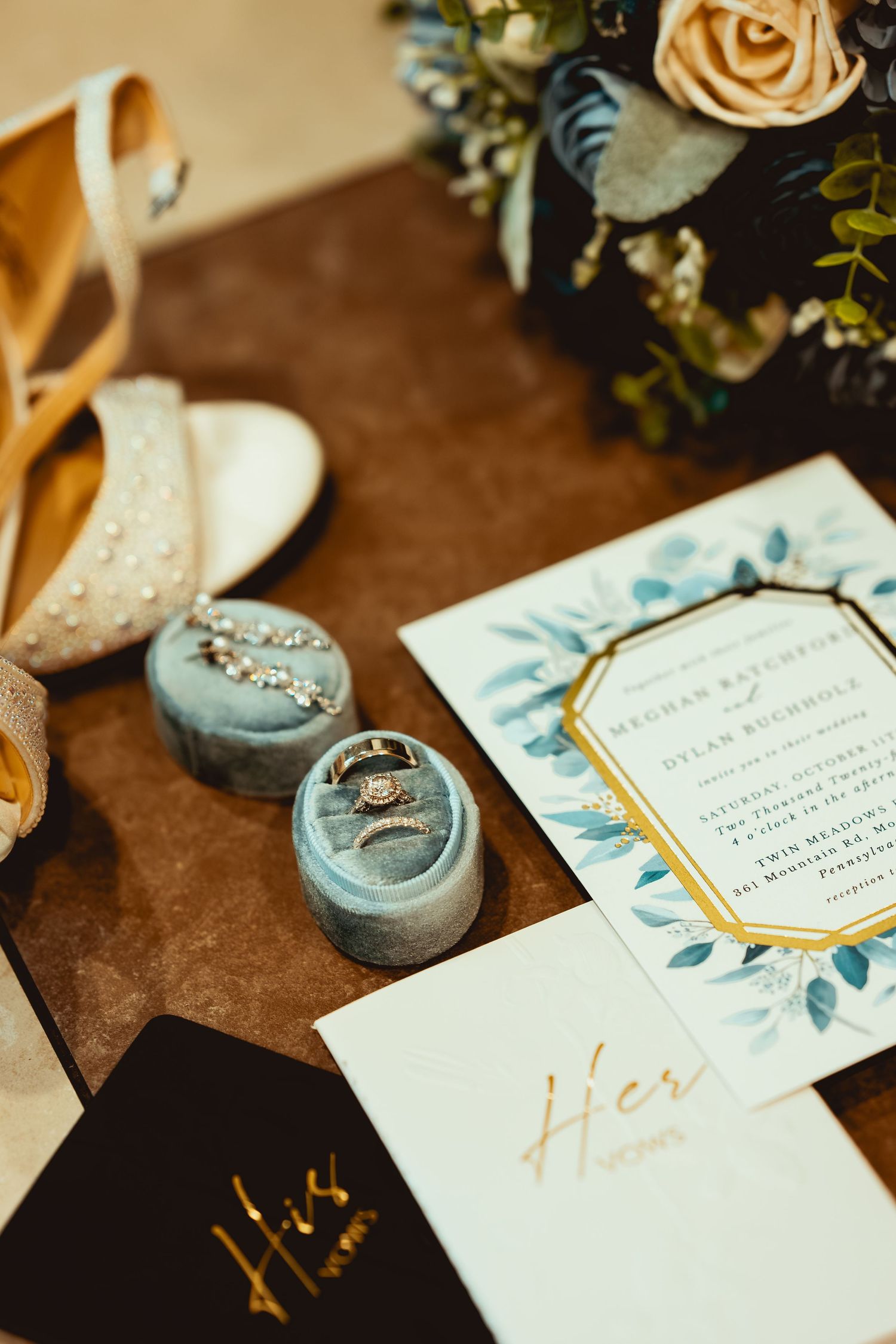 Wedding flat lay with rings in blue velvet box, invitation, bridal shoes, bouquet, and His and Hers vow books.