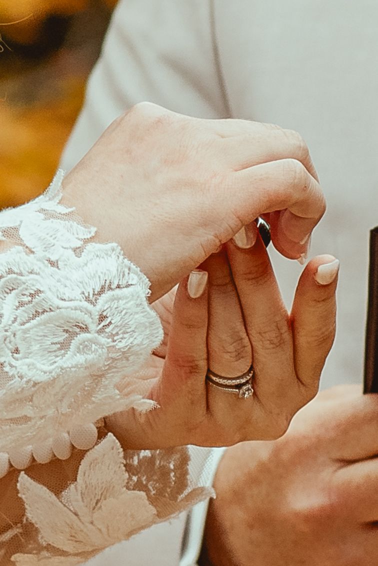Close-up of a bride in lace dress receiving a diamond wedding ring on her finger during a ceremony.