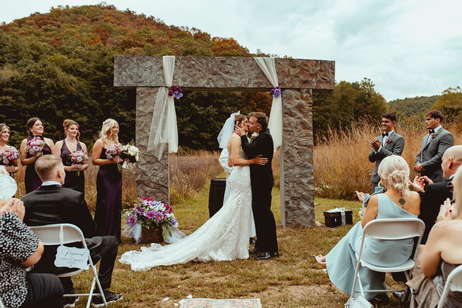 Outdoor wedding ceremony taking place at a scenic mountainside stone structure with guests gathered around.