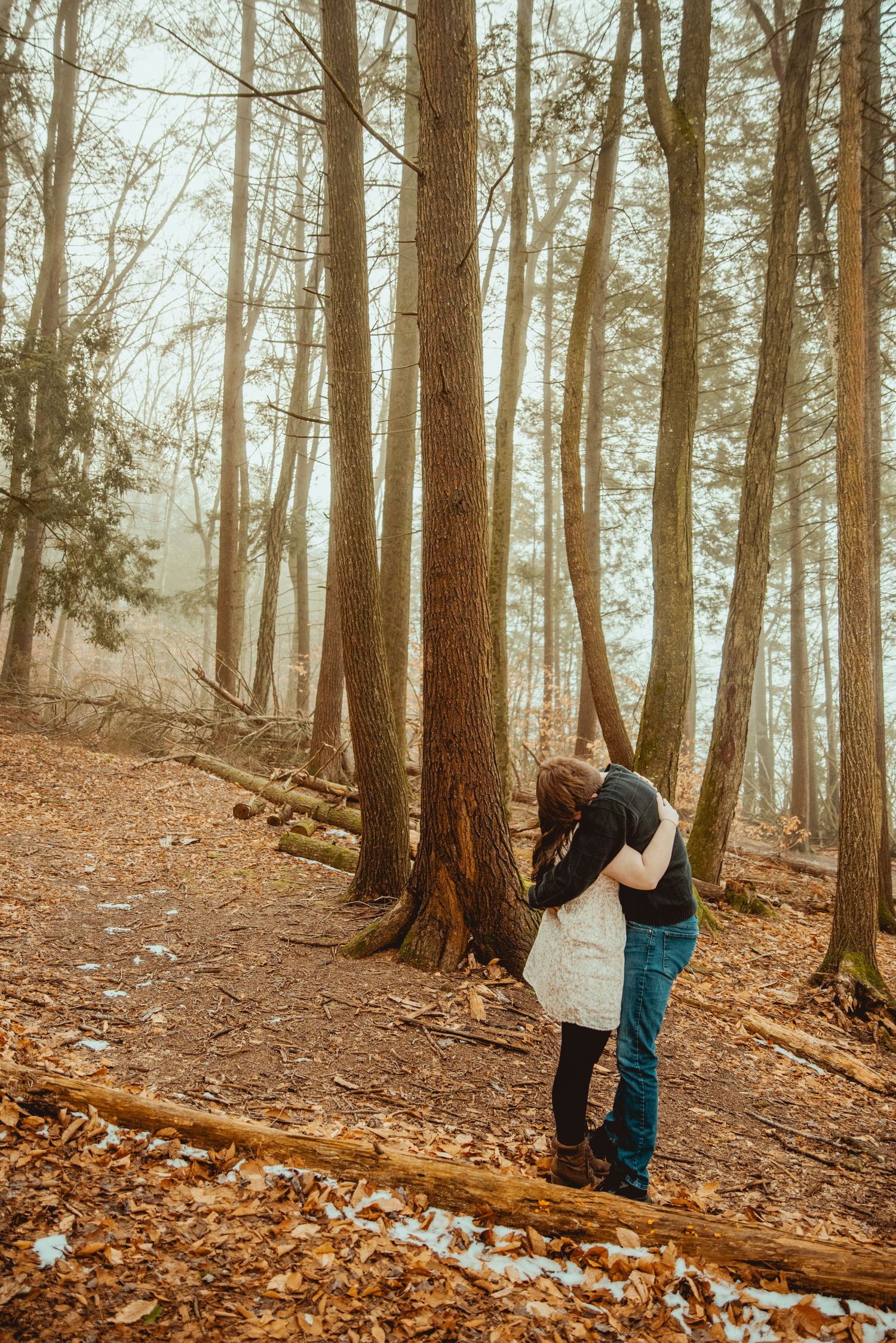 Two people stand together among tall trees in a misty autumn forest with fallen leaves covering the ground.