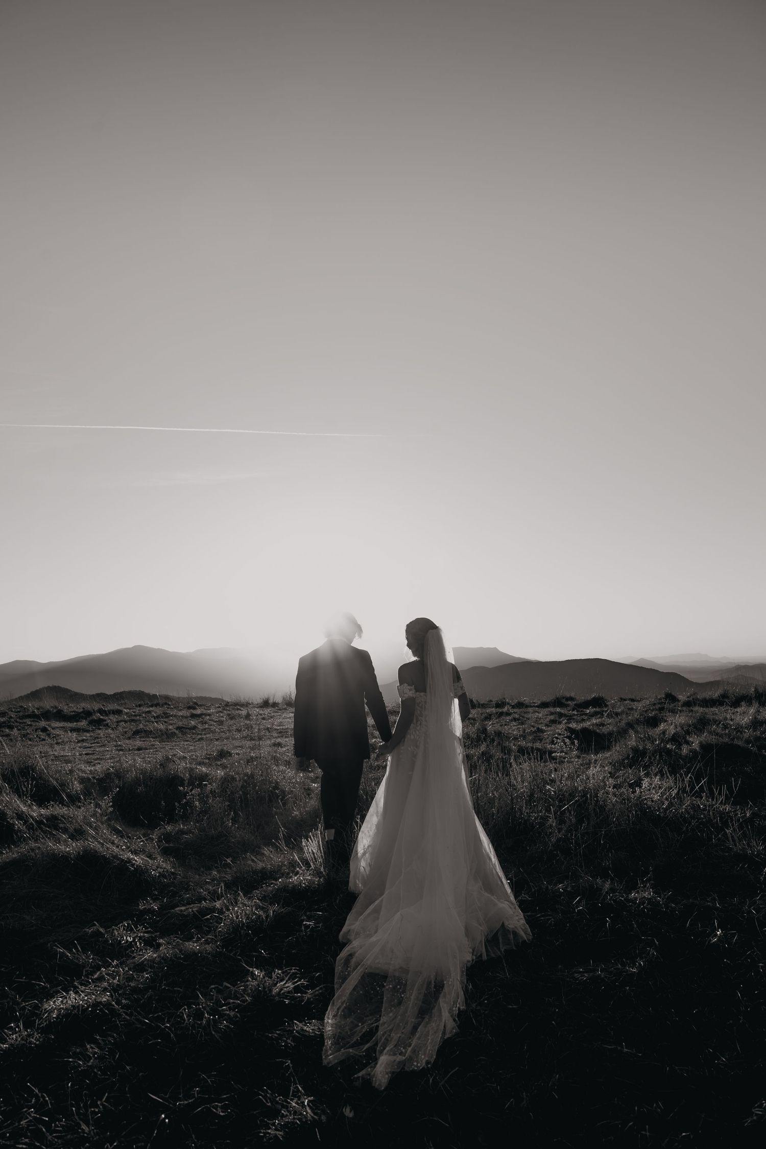 Black and white wedding photo of couple walking through mountain field at sunset.