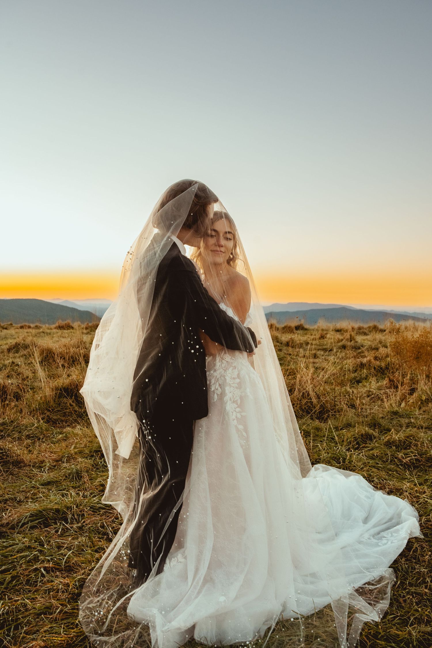 Couple embraces in wedding attire during golden sunset in rustic field with mountains in background.