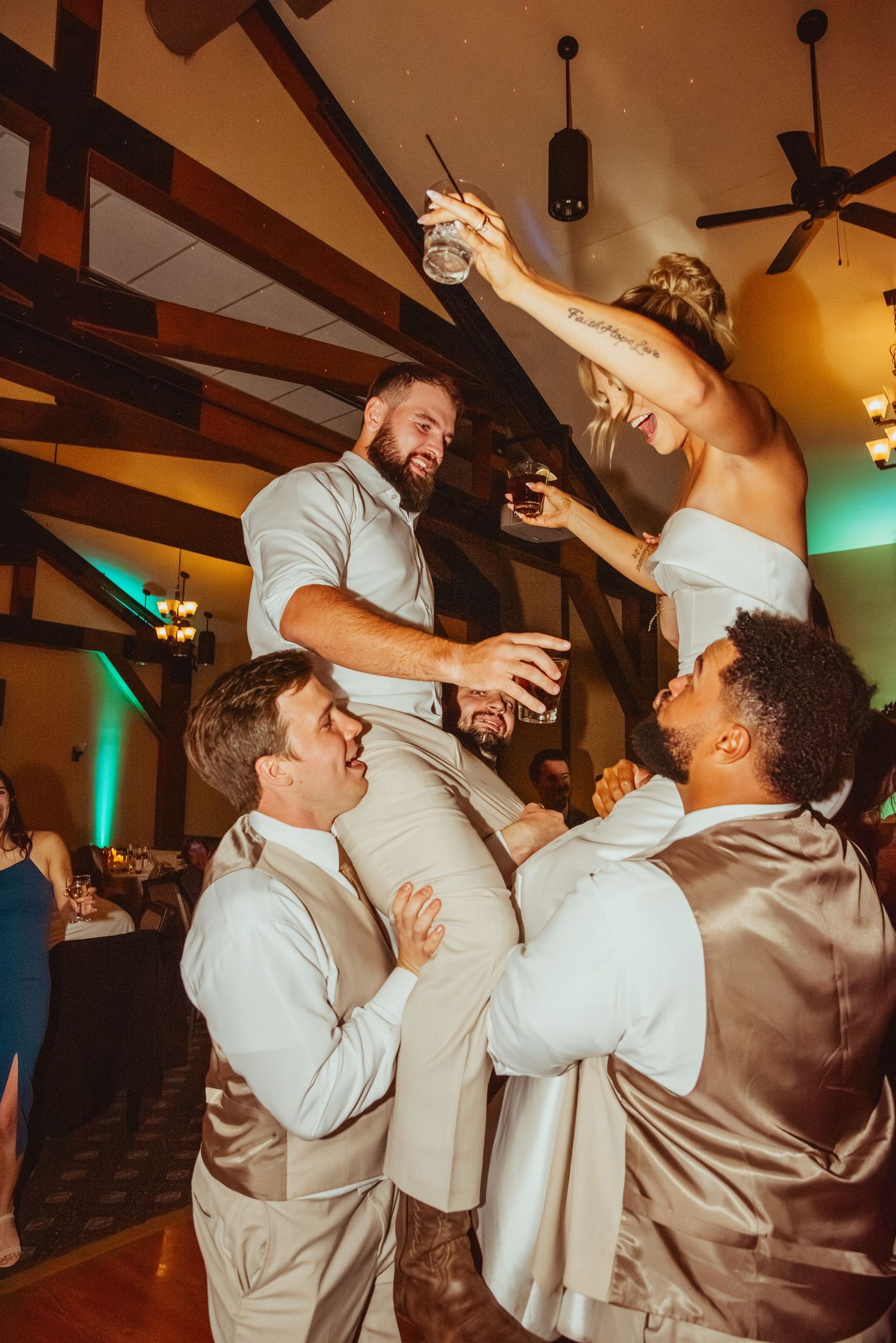 Wedding party guests dancing and celebrating energetically on an indoor dance floor with wooden beams overhead.