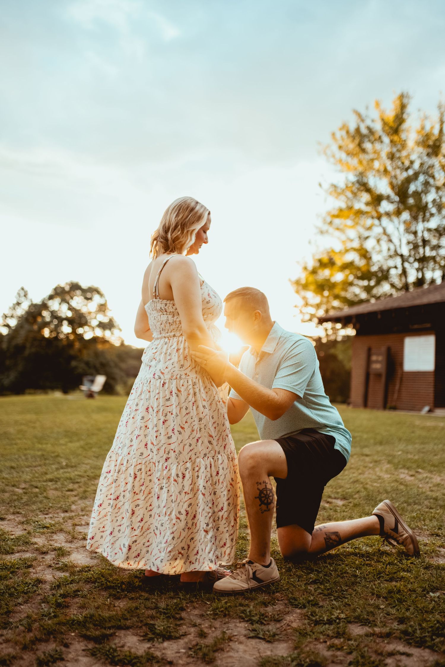 Romantic sunset proposal scene with couple in a grassy field near a rustic building.