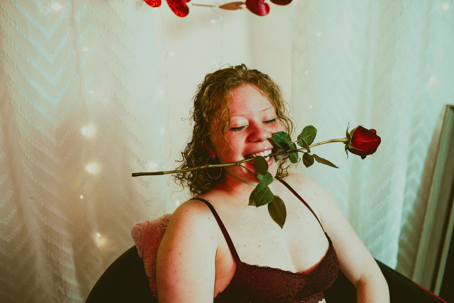 A red rose is held between teeth against a soft curtained background in a dreamy vintage-style photograph.