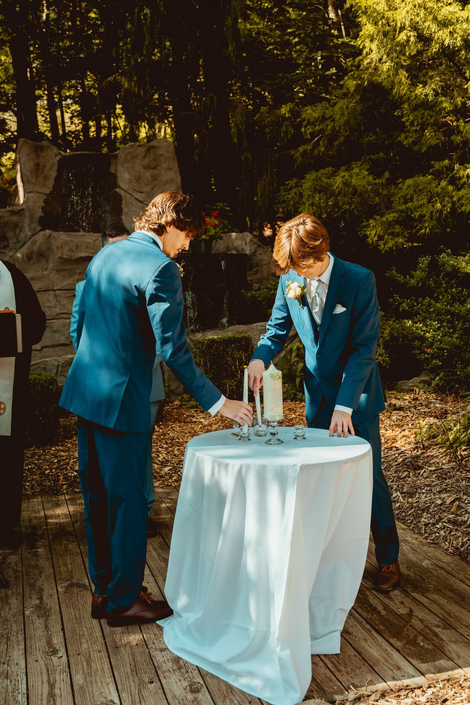 Wedding party in teal blue suits signing documents at a white-clothed table on a wooden deck surrounded by trees.