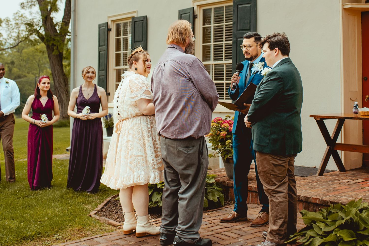 An intimate outdoor wedding ceremony takes place on a green lawn in front of a historic house with white columns.