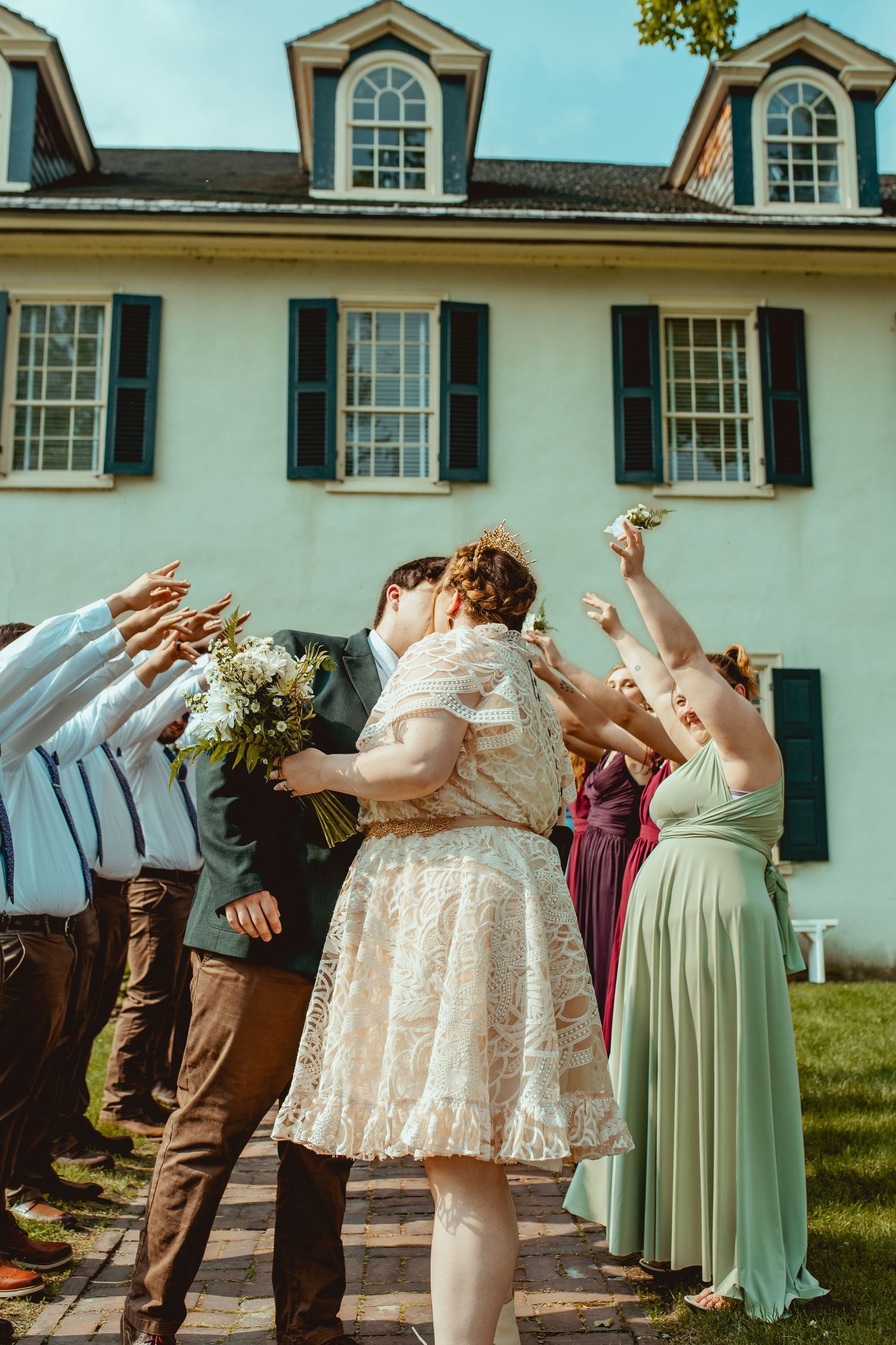 Wedding party members perform synchronized dance moves on a lawn in front of a white colonial style building.