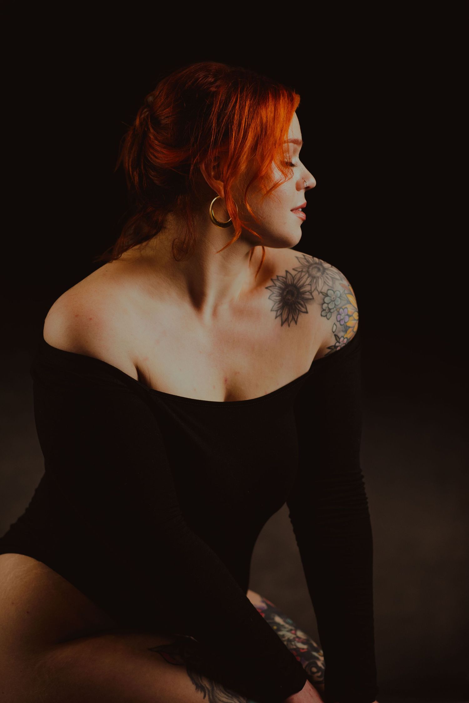 Redhead woman in black off-shoulder top with floral shoulder tattoo poses against dark background.