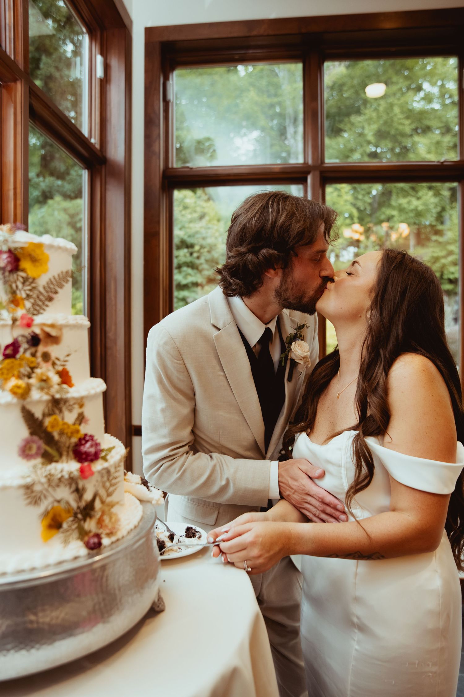 Bride and groom share a kiss while cutting their floral-decorated wedding cake near a window.