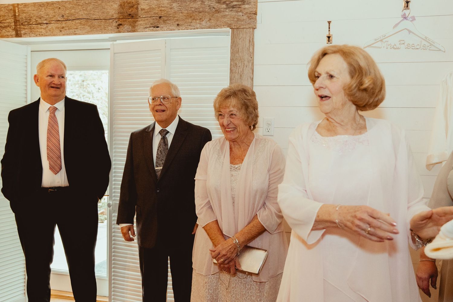 Elegantly dressed wedding guests smiling and conversing in a bright rustic venue.