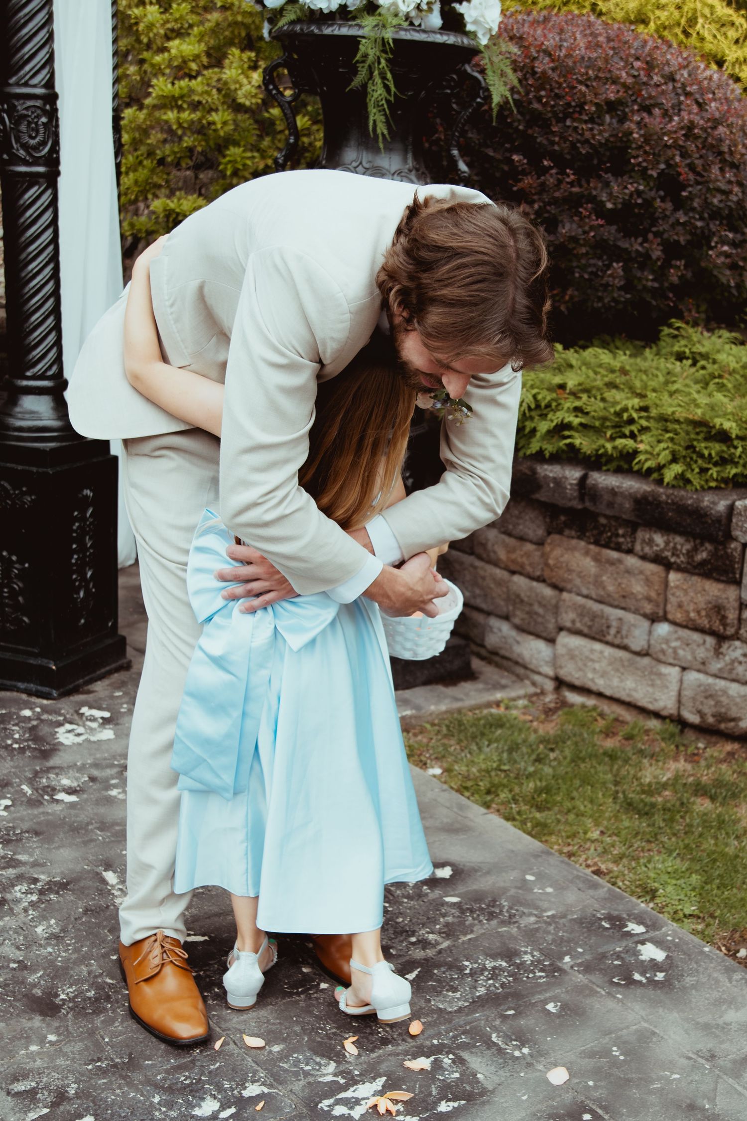 Groom in beige suit bending down to hug bride in light blue dress outdoors at wedding venue.