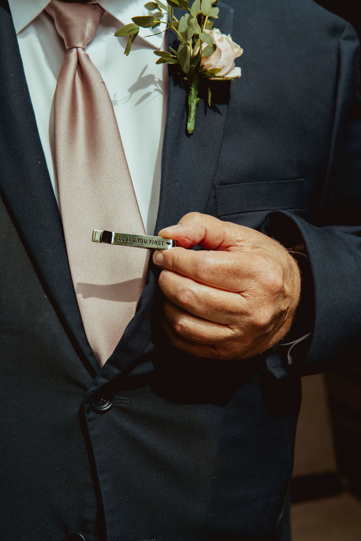 Groom in navy suit with pink tie holds engraved tie clip near boutonniere at wedding.