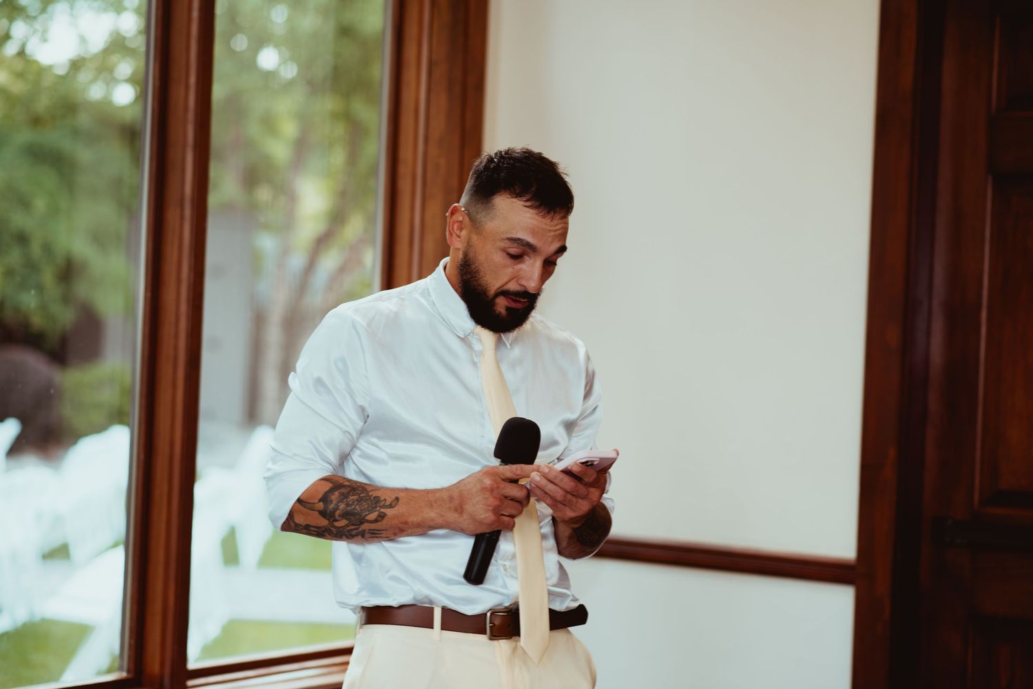 Tattooed man in white shirt holding microphone and notes while delivering a wedding speech indoors.