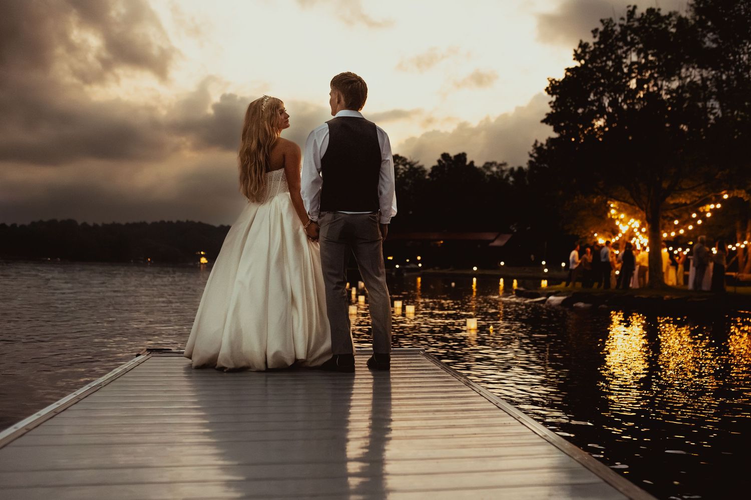 Bride and groom holding hands on a lakeside dock at sunset with string lights glowing in the background.