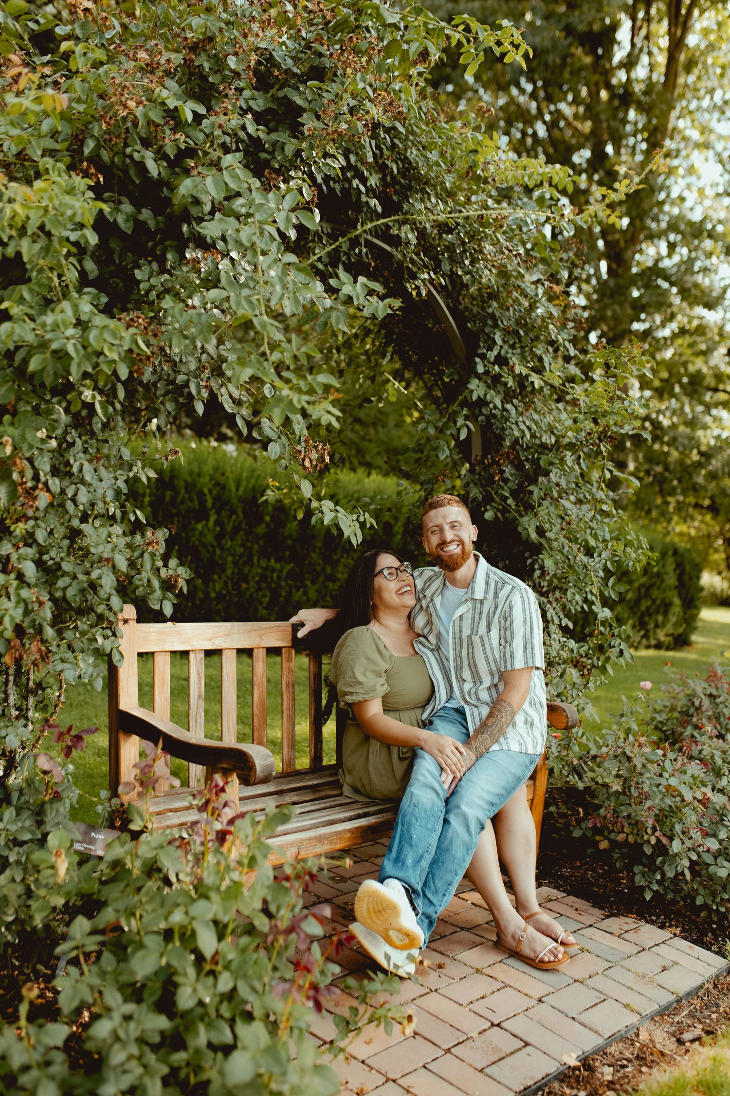A smiling couple sits closely on a wooden garden bench surrounded by lush green trees and shrubs.