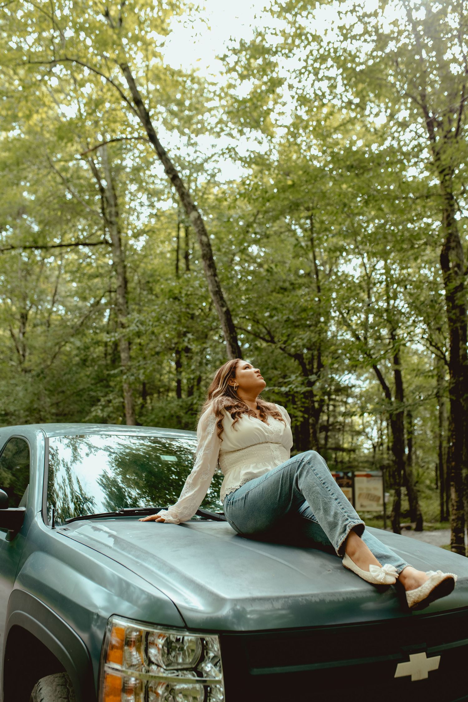 Woman in white sweater and jeans sitting on hood of Chevrolet truck surrounded by lush green forest trees.