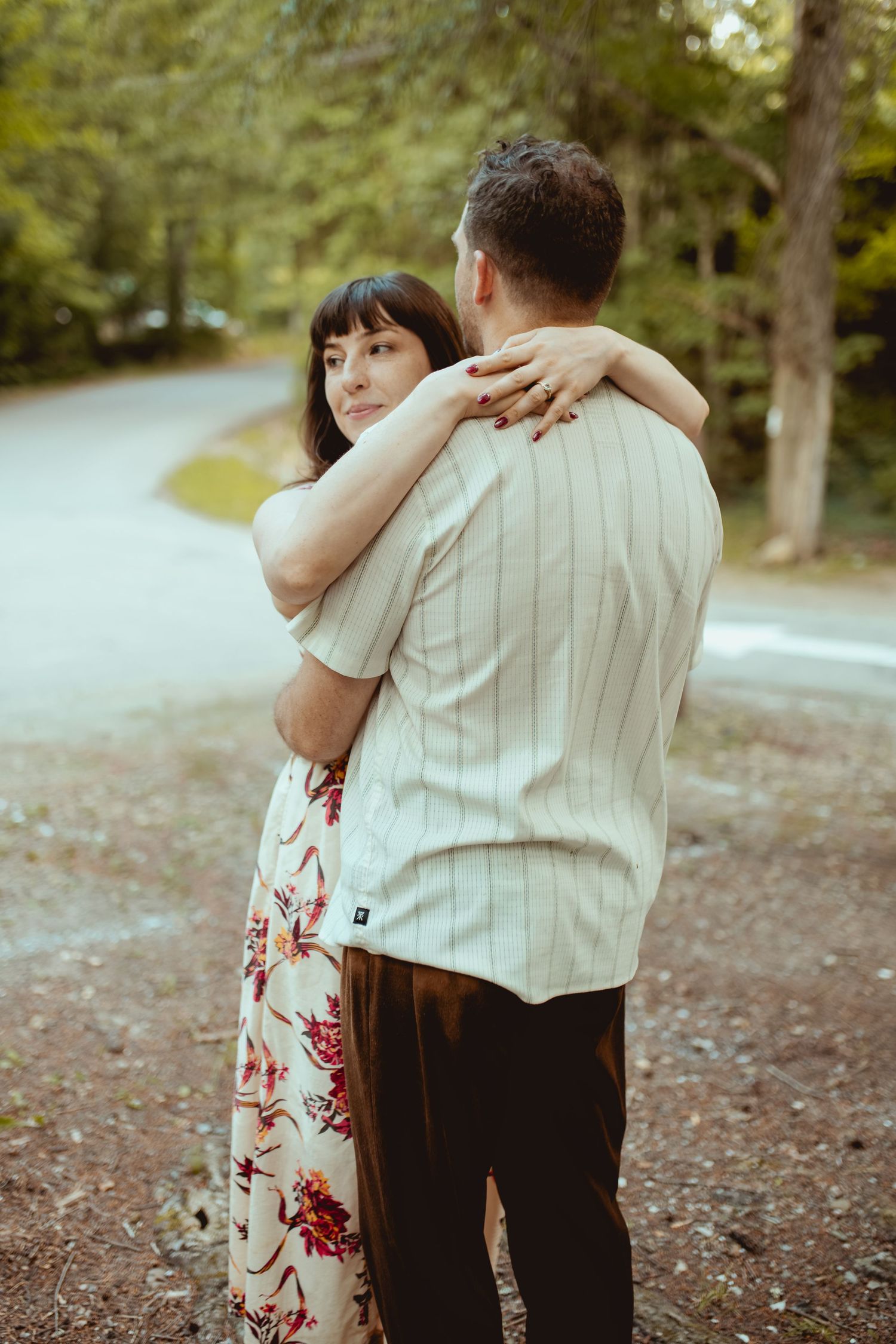 A couple embraces outdoors near a forest road, woman in floral dress hugging man in striped shirt.