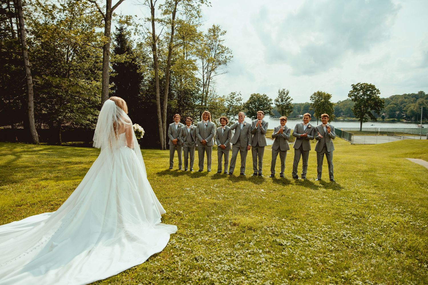 Bride in white gown with long veil stands before groomsmen in gray suits on green lawn near lake.