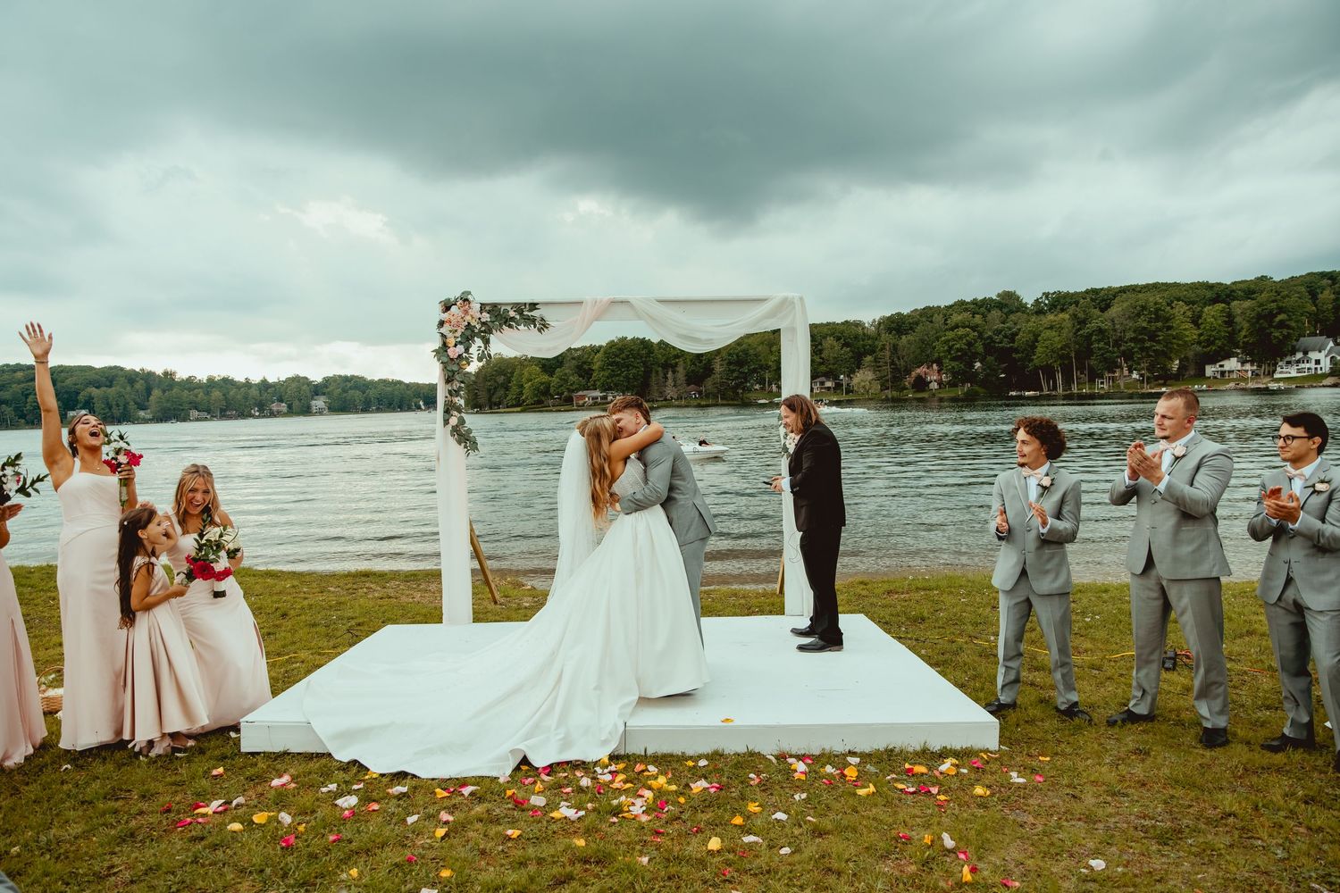 Couple shares first kiss at outdoor lakeside wedding ceremony with white arch and cheering guests.