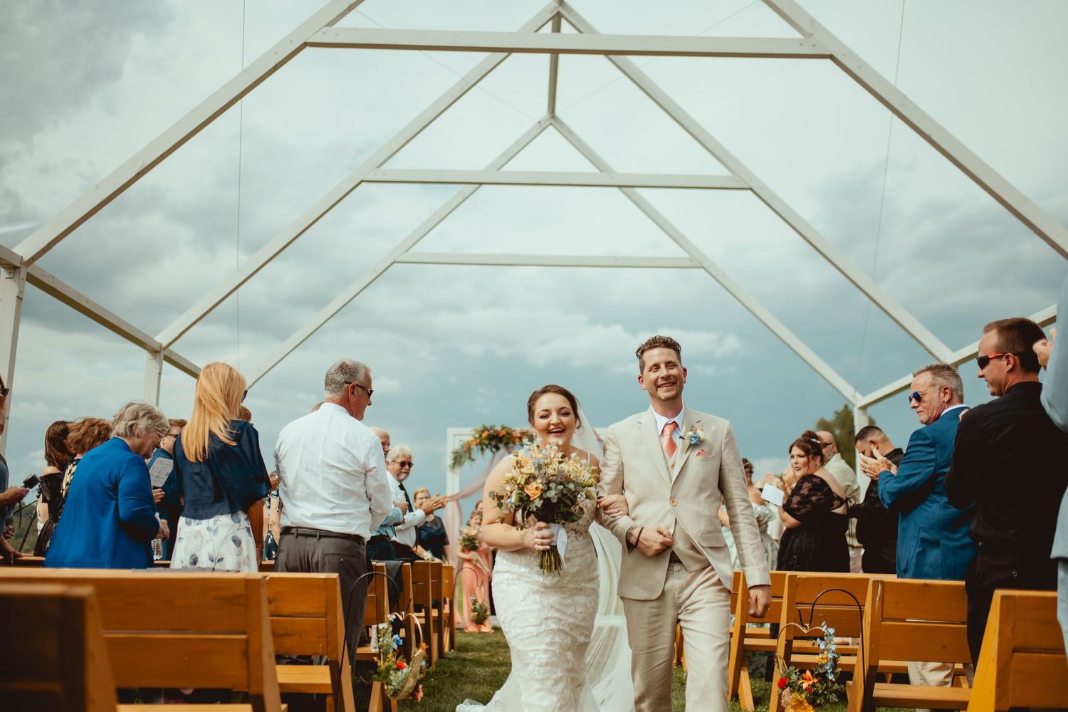 Bride and groom walk down the aisle under a clear tent at an outdoor wedding ceremony.