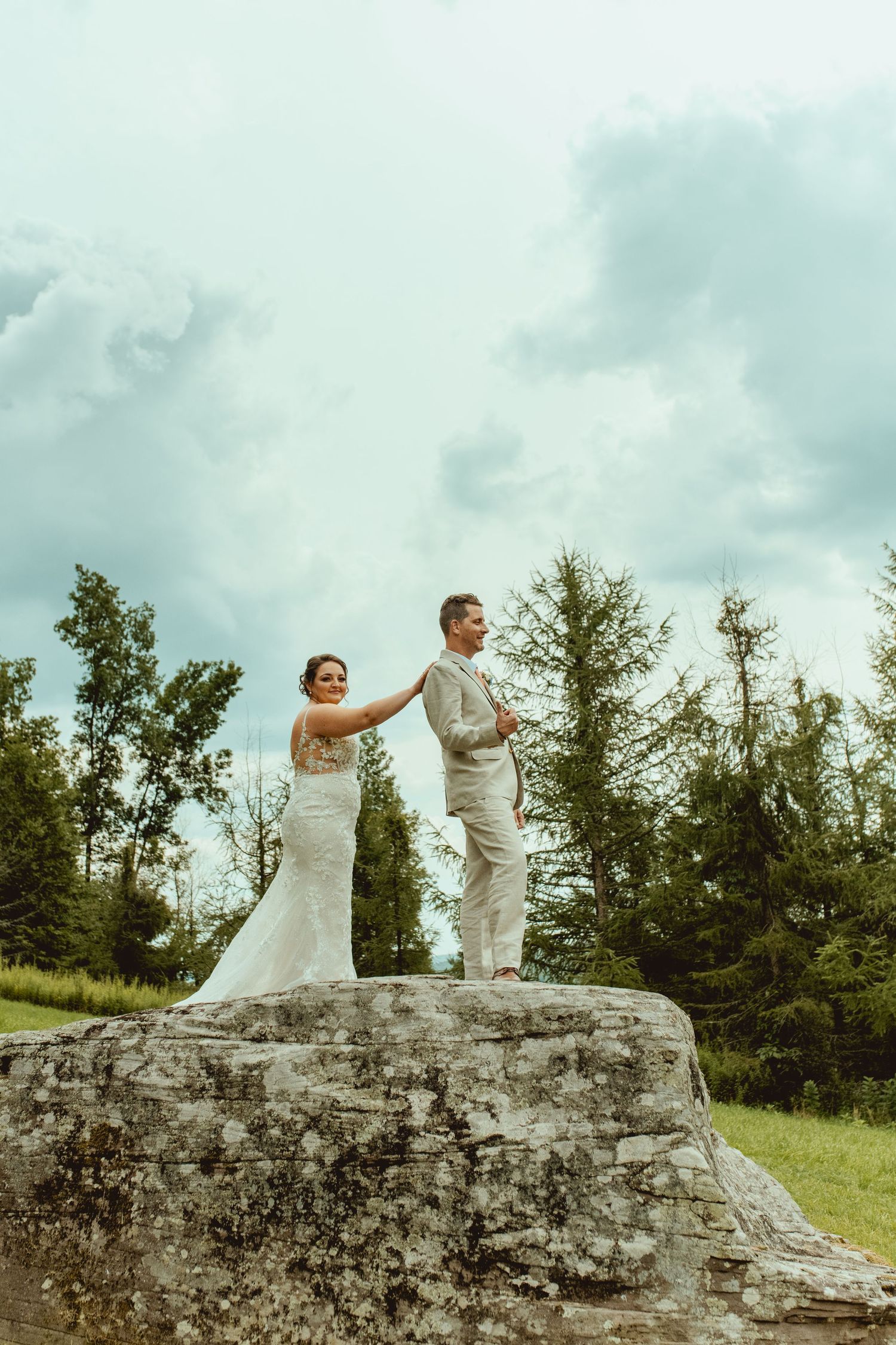Bride and groom stand atop a stone rock surrounded by lush green trees under a cloudy sky.