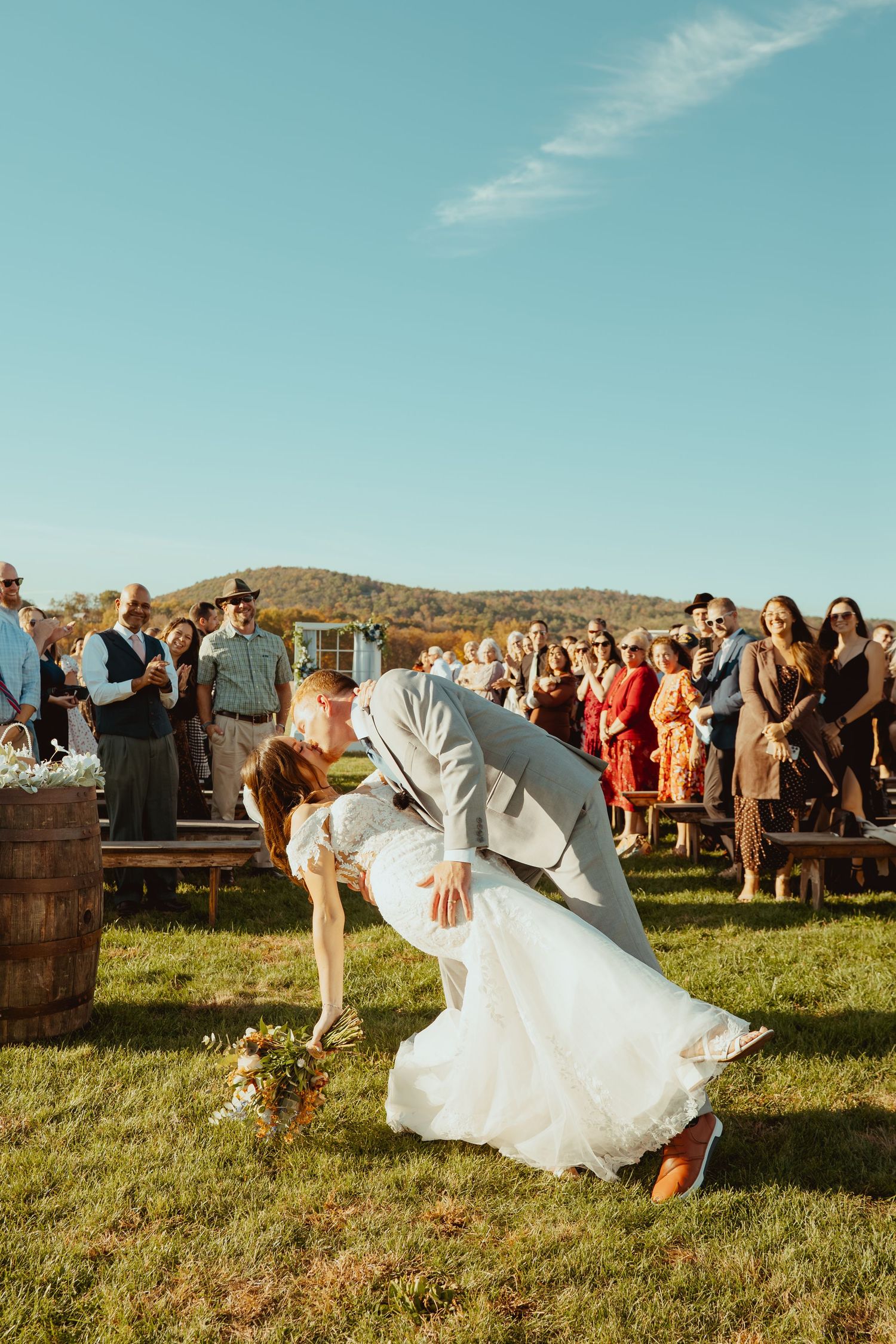 Newlyweds perform a playful dip on grassy field during outdoor wedding with guests watching in background.