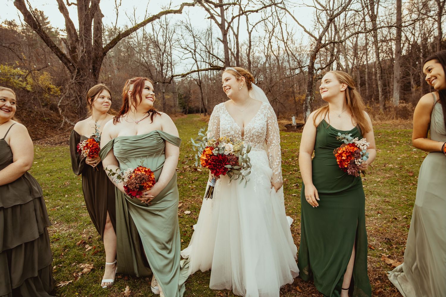 Wedding party members in green and gray dresses pose together holding red bouquets in an autumn outdoor setting.