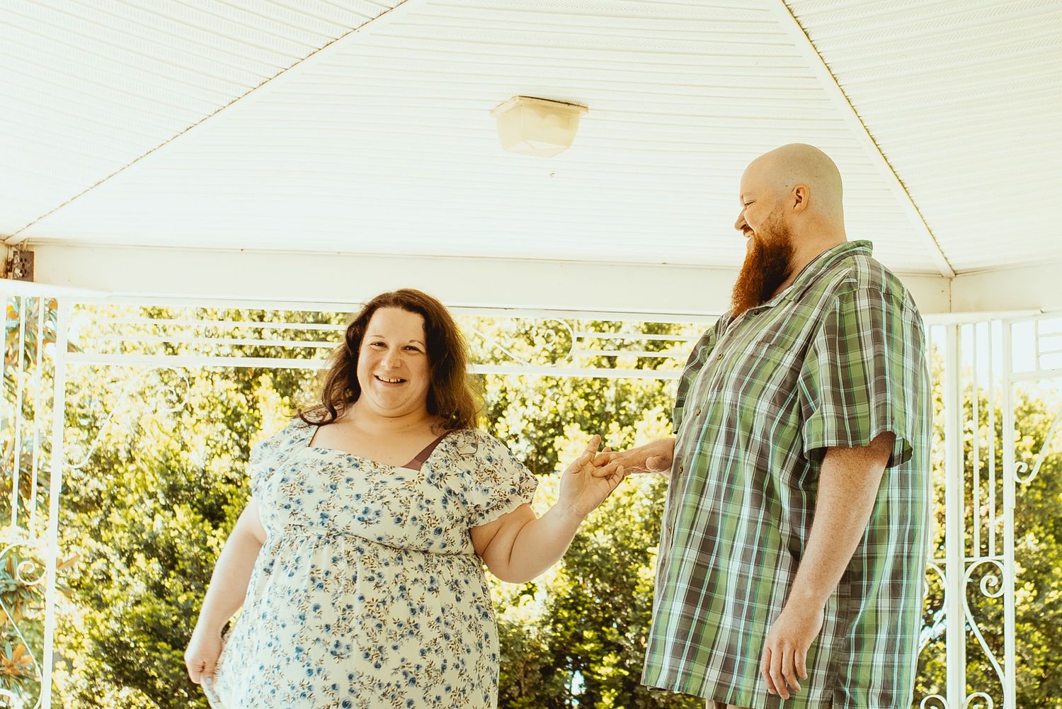 A smiling couple holds hands outdoors under a white pergola, woman in floral dress, man in plaid shirt with beard.