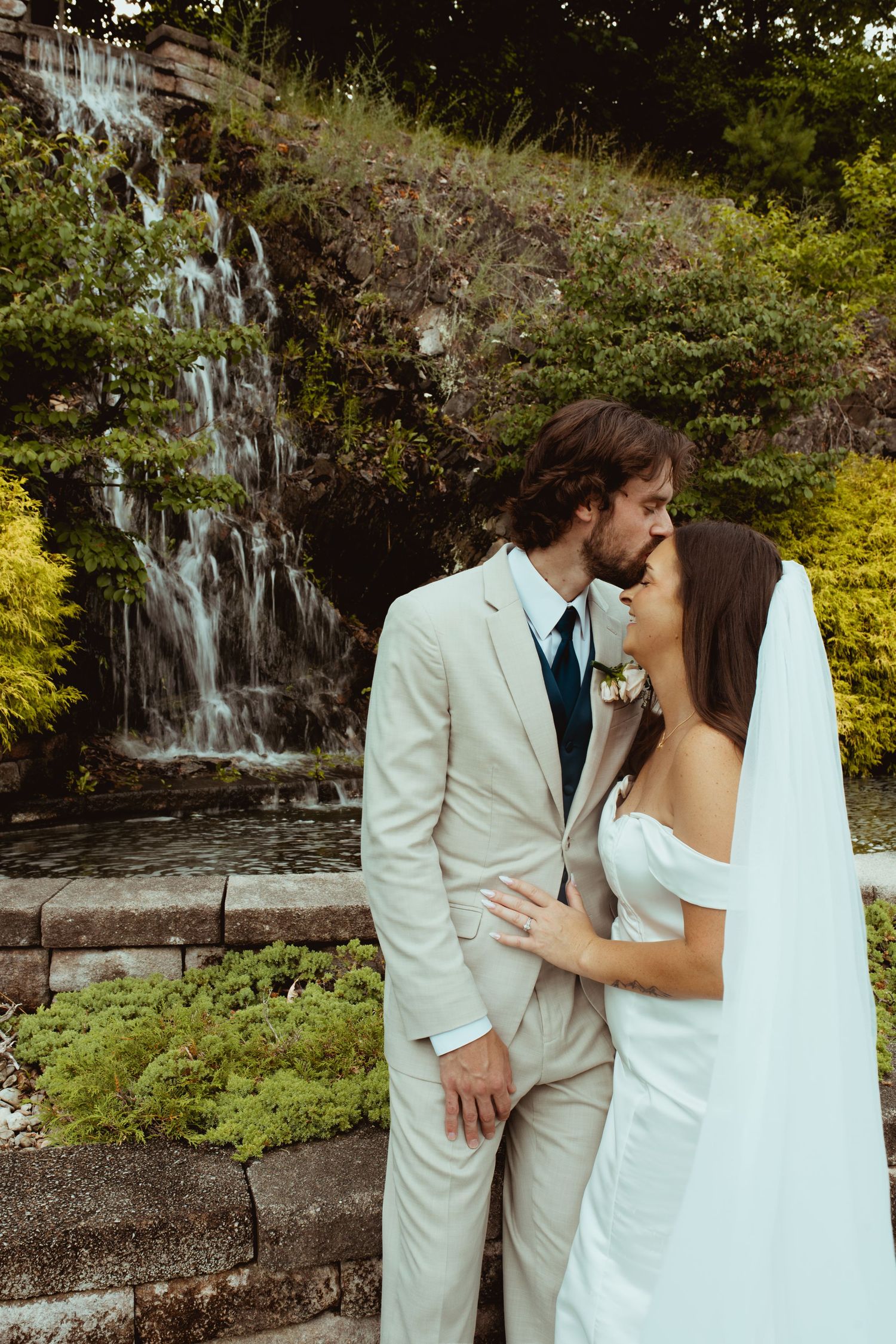 Bride and groom share a romantic moment in front of a waterfall, dressed in white wedding attire outdoors.
