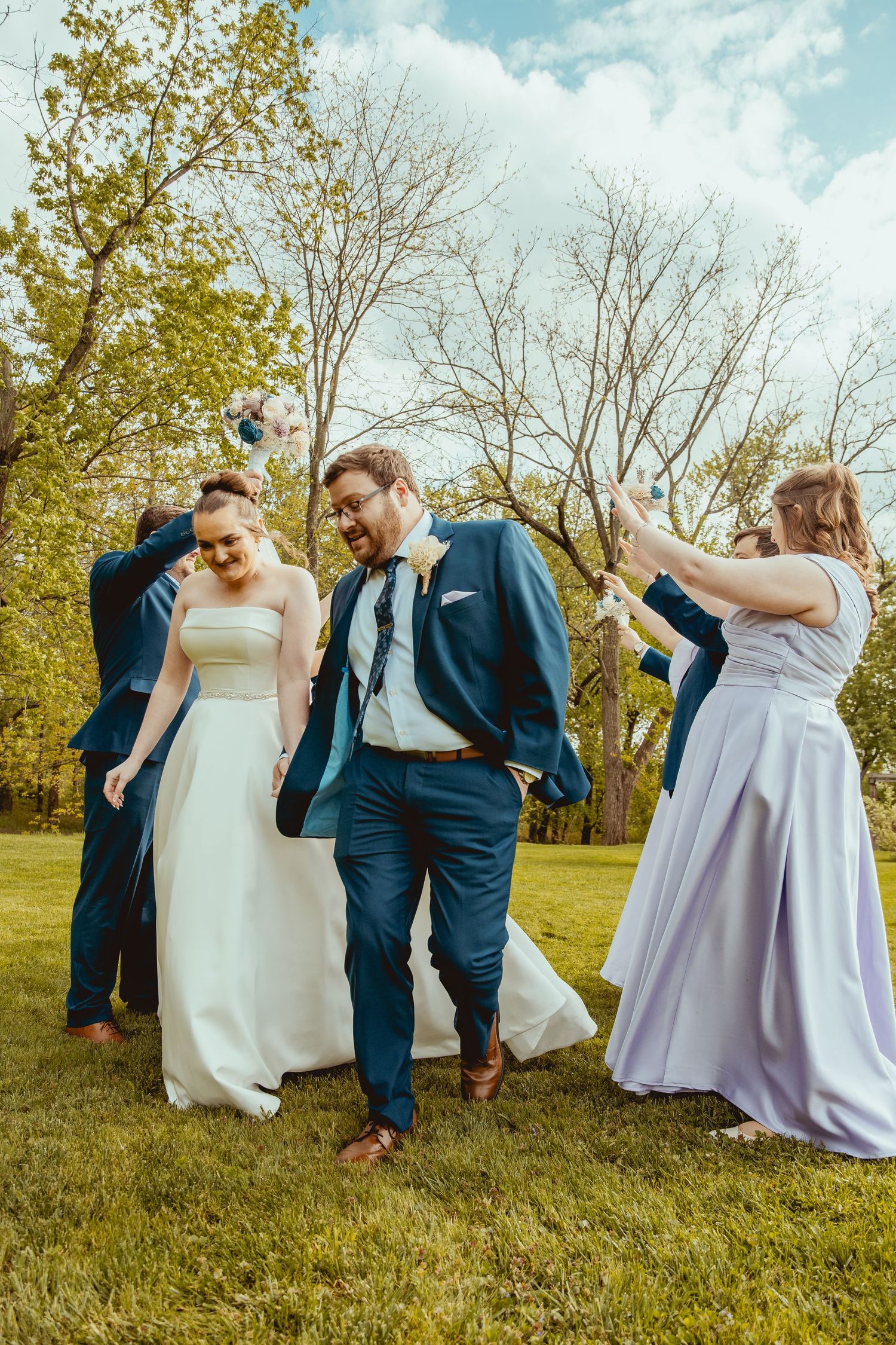 Joyful wedding party celebrating outdoors on green grass with trees, bride in white gown and groom in navy suit.