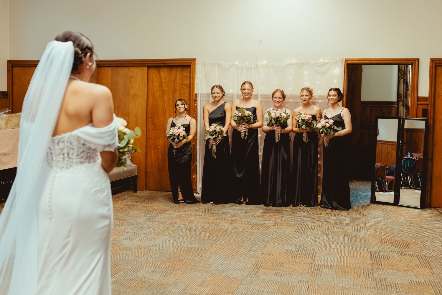 Bride and bridesmaids in black dresses pose together in a line against a stone wall.