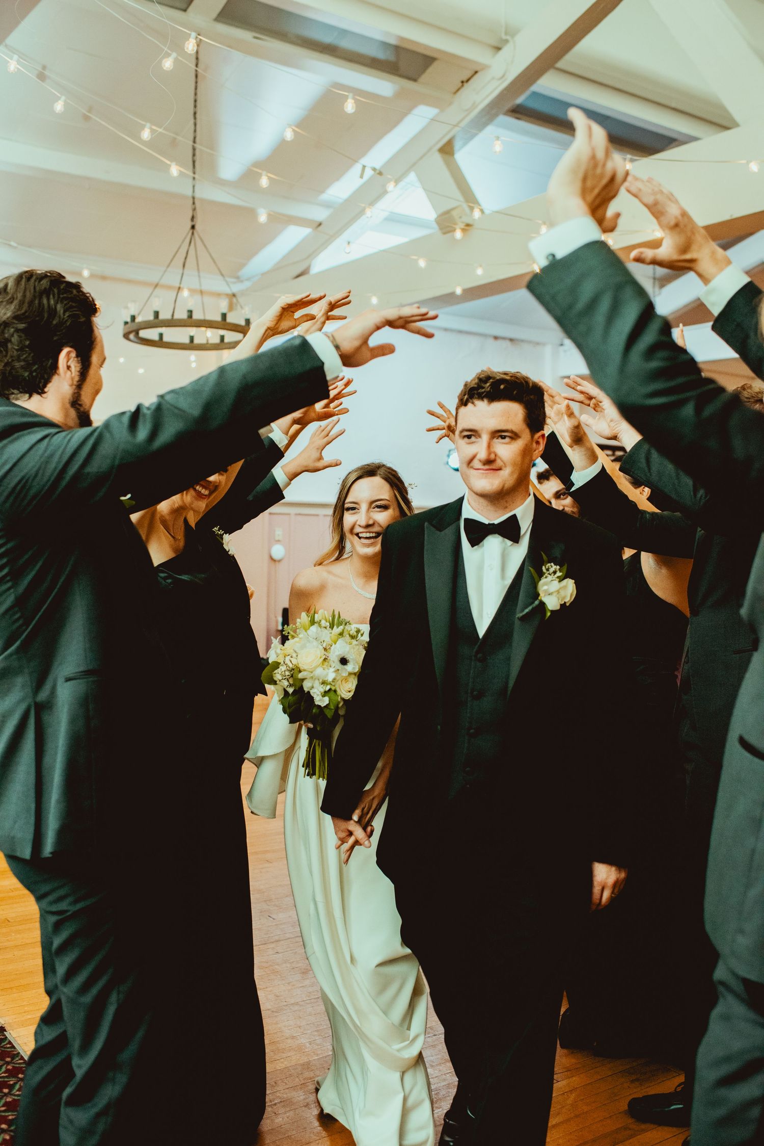 Wedding party members in dark suits celebrate with raised arms in a reception hall.