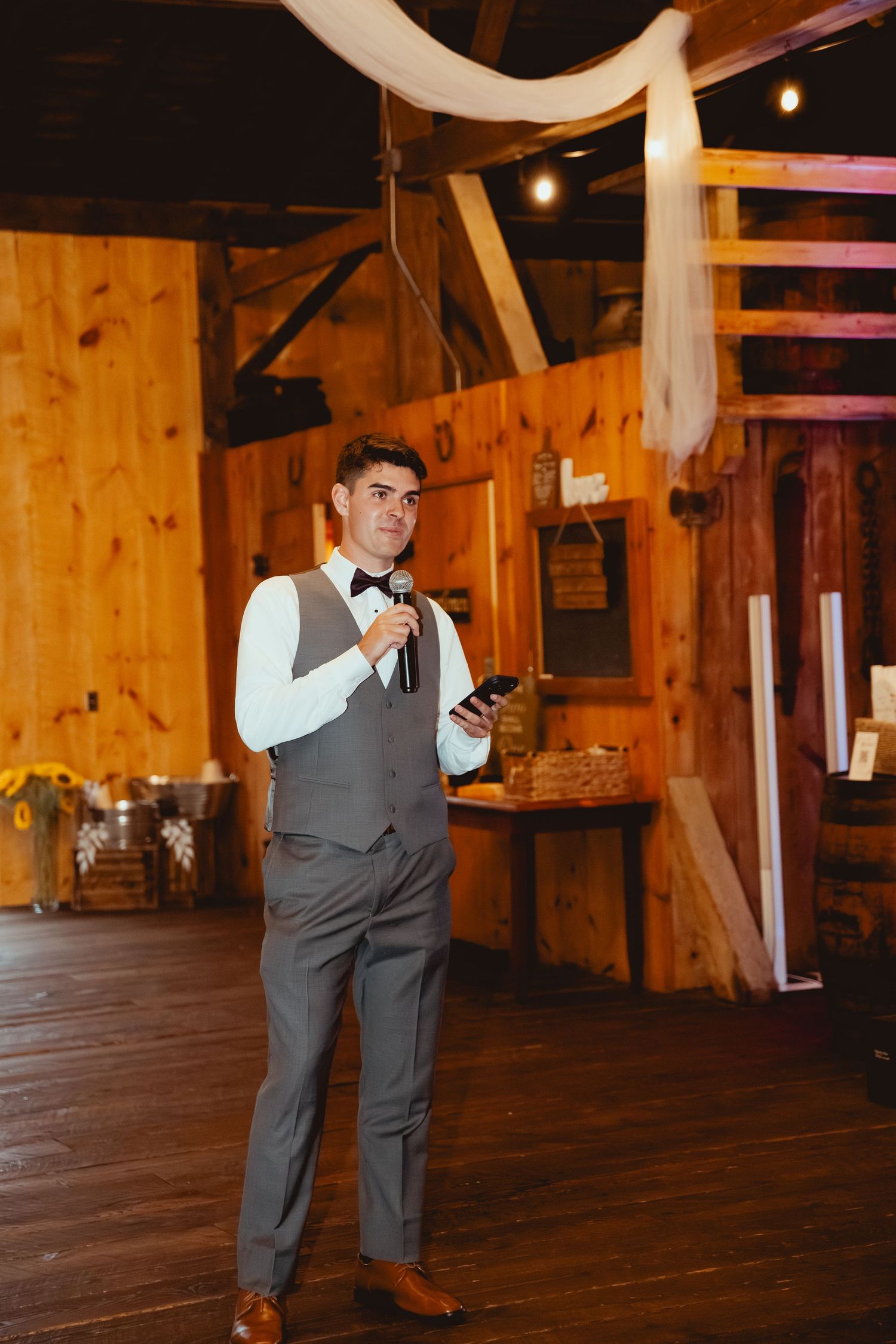 Groomsman in gray vest checks phone while standing in rustic wooden barn venue with warm lighting.