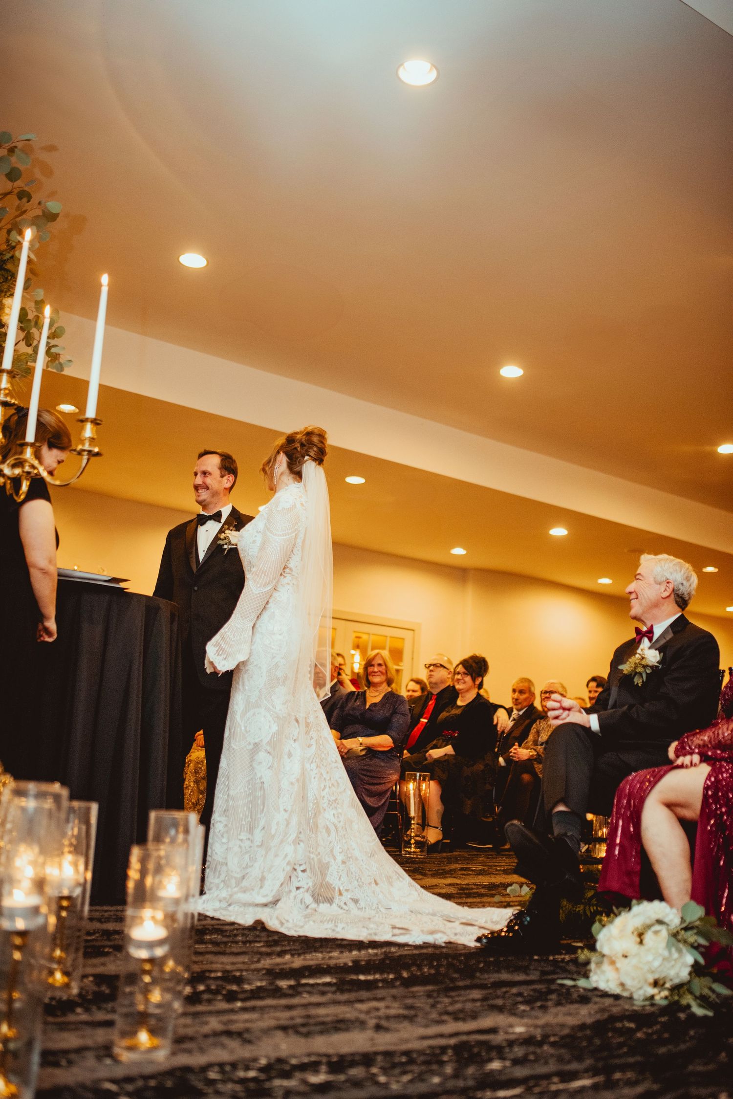 Bride in a lace gown with a long veil faces her groom at the altar as seated wedding guests watch during an indoor ceremony with candles.